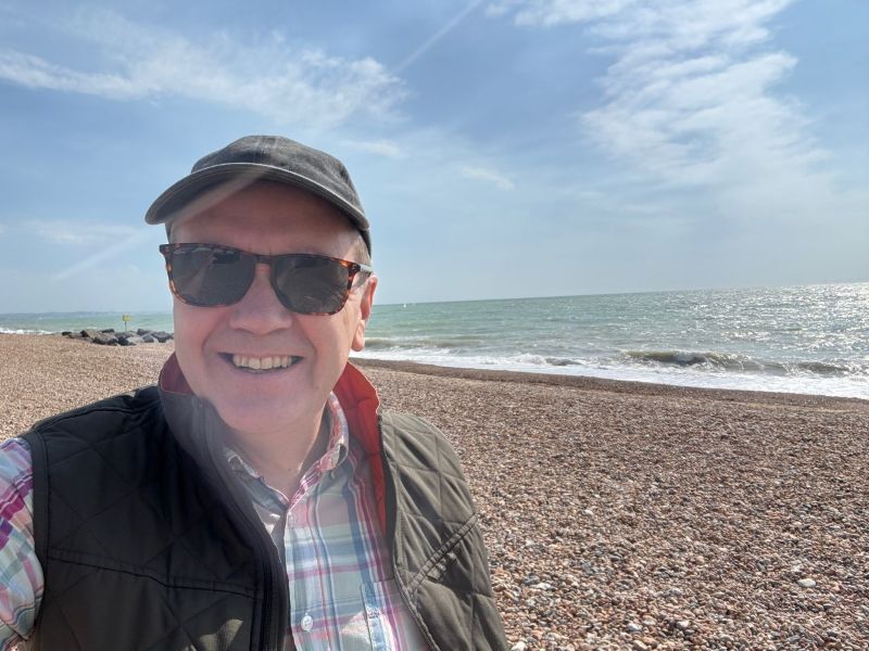 A man wearing sunglasses, a black cap, and a checked shirt with a waistcoat smiles at the camera whilst standing on a pebbly beach with waves and a partly cloudy sky in the background.