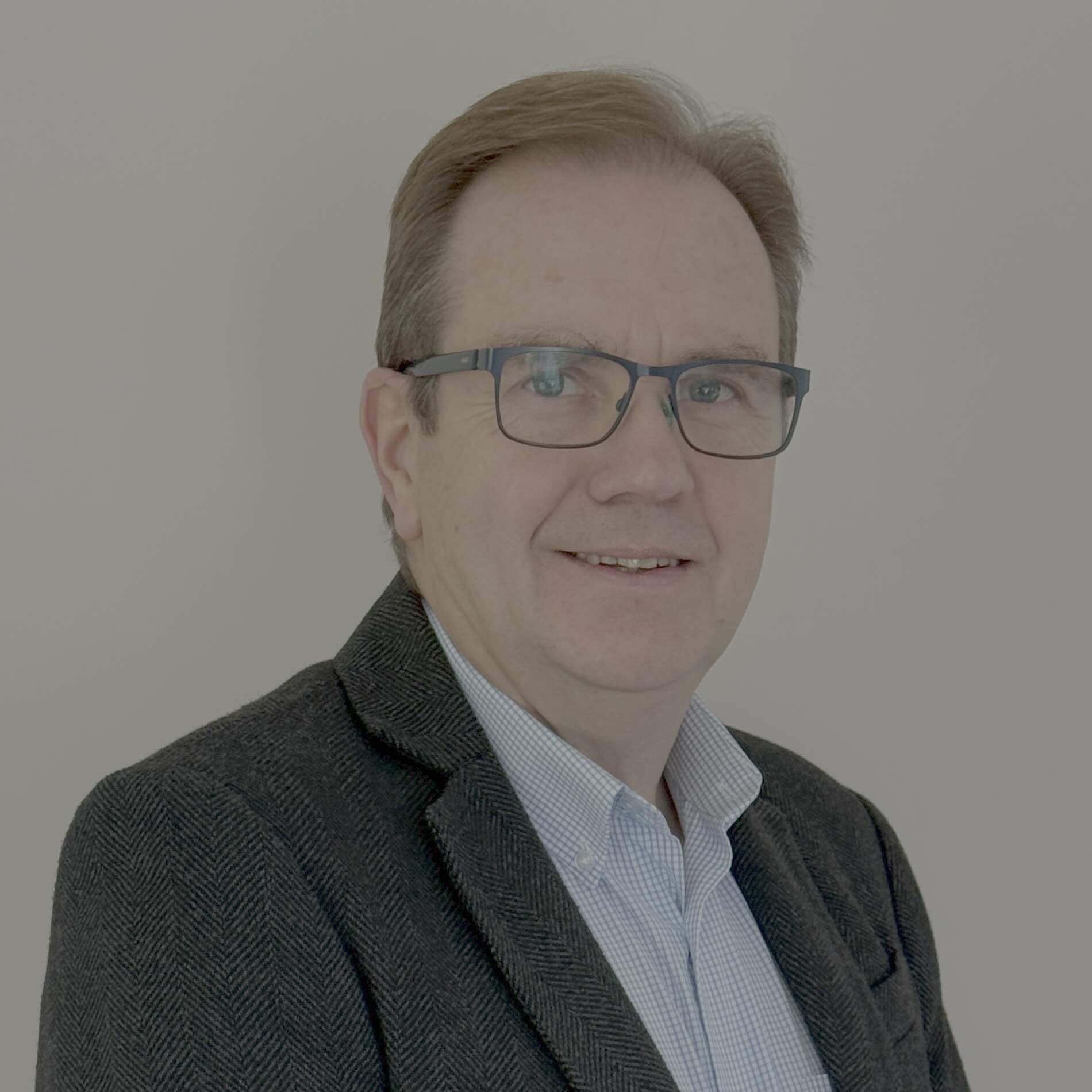 A middle-aged man with short light brown hair and glasses, wearing a dark blazer over a light blue shirt, smiling and posing in front of a plain white background.